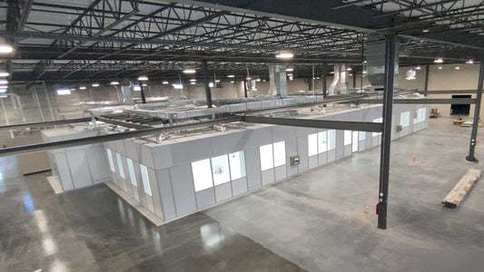 A large modular 7500 square ft ISO 8 Cleanroom with white panel walls and multiple windows, situated inside a spacious industrial warehouse with a high steel truss ceiling and concrete floor.