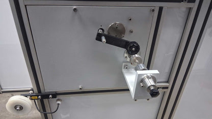 A close-up of a mmbt 96 carrier horizontal bobbin winder mounted on a gray panel, featuring a black and silver arm, a white mounting block, and a small white wheel on the left.