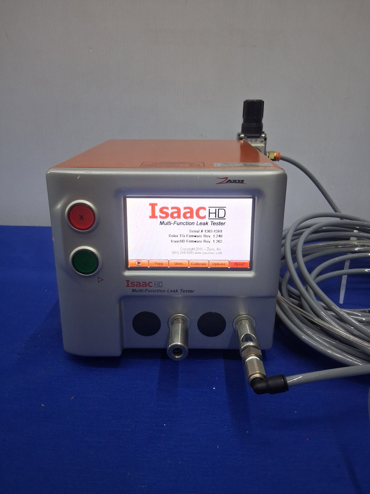 An Isaac HD Multi Function Lead Tester device with a gray casing, orange top cover, red and green buttons, and attached gray cables, set against a blue surface and gray background.
