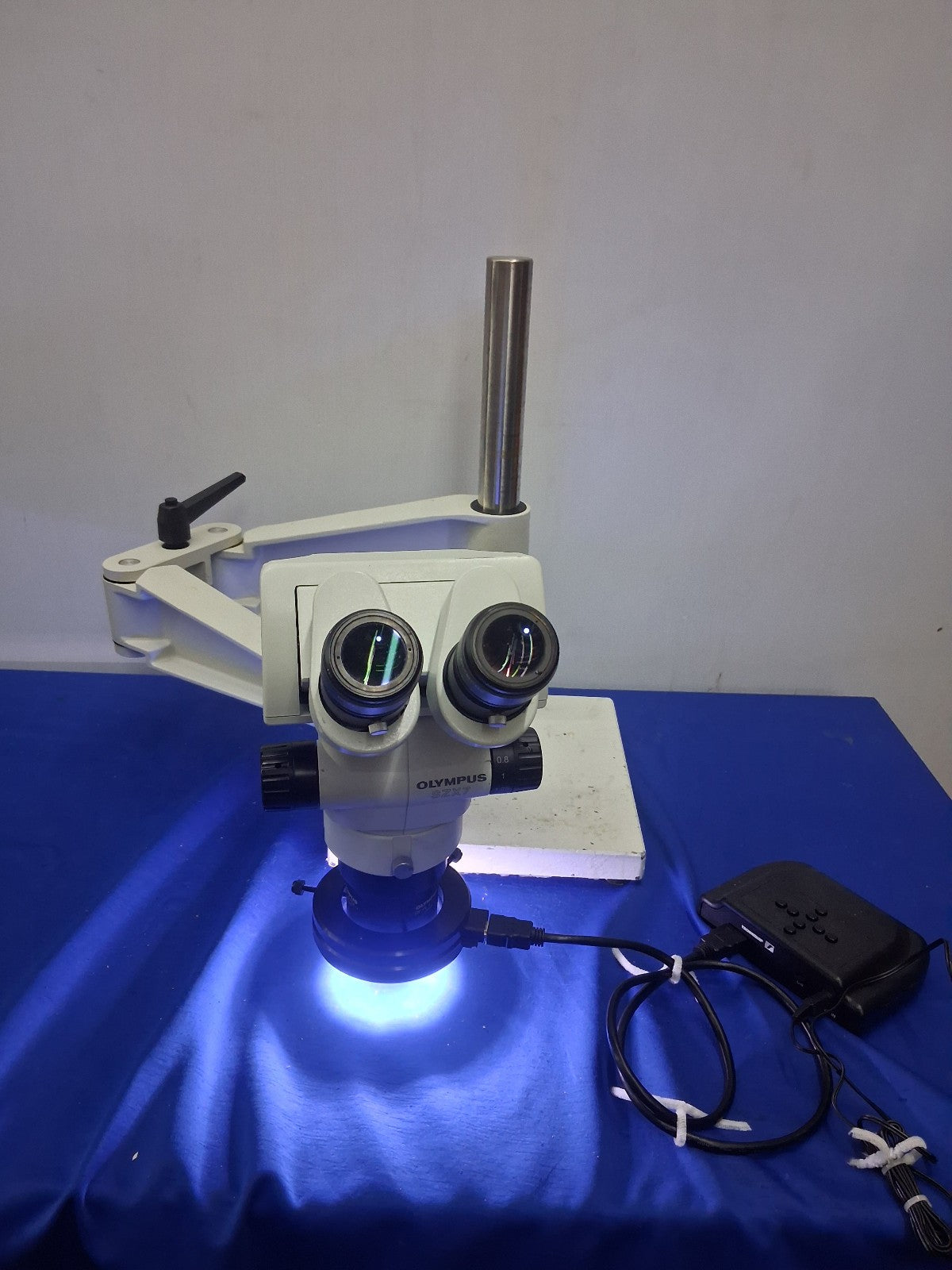 An Olympus binocular microscope with an integrated light source, shown on a blue cloth-covered table with its power adapter nearby.