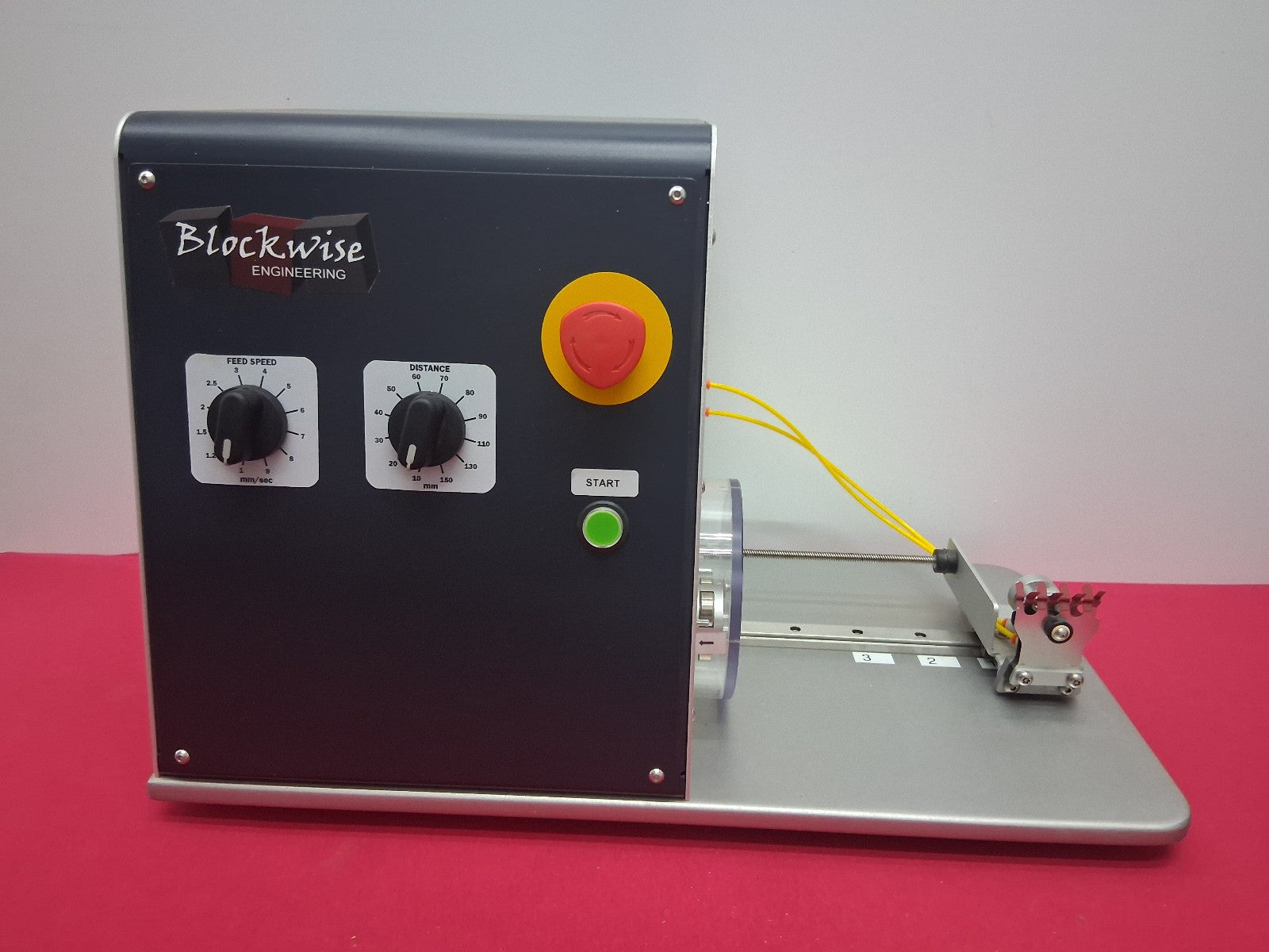 A Blockwise Engineering industrial testing machine with a black control panel, silver base, yellow and green wires, and a metal sample holder, set against a plain gray background and red surface.