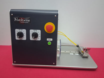 A Blockwise Engineering industrial testing machine with a black control panel, silver base, yellow and green wires, and a metal sample holder, set against a plain gray background and red surface.