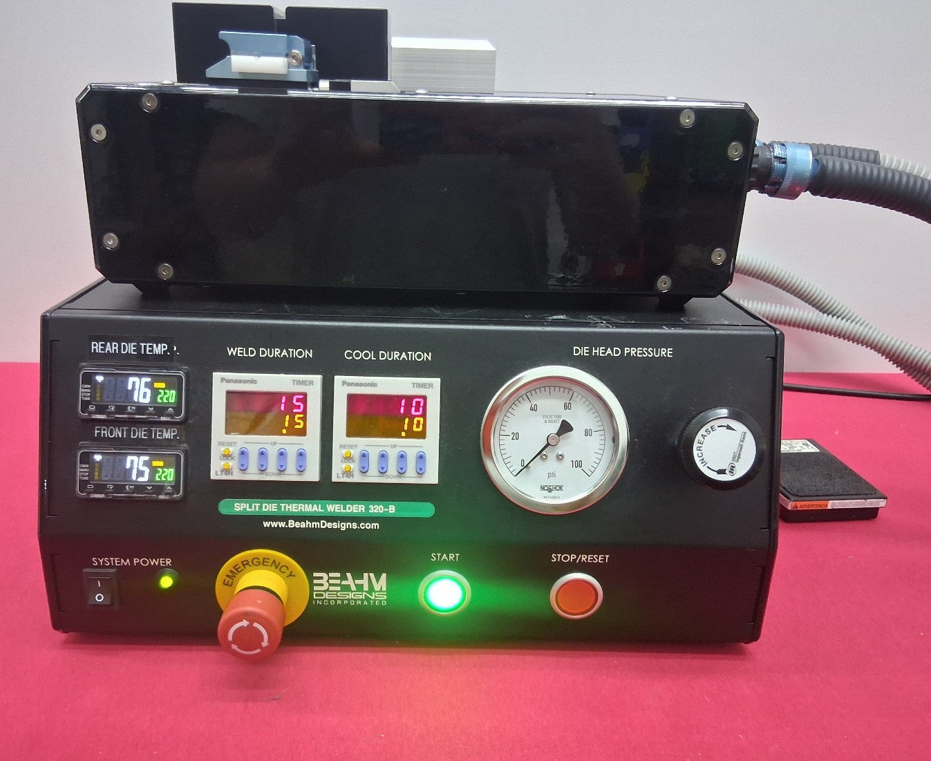 A black industrial die cutting machine with dual digital temperature displays, gauges, and control buttons, shown on a pink surface with a black frame or box mounted above it.